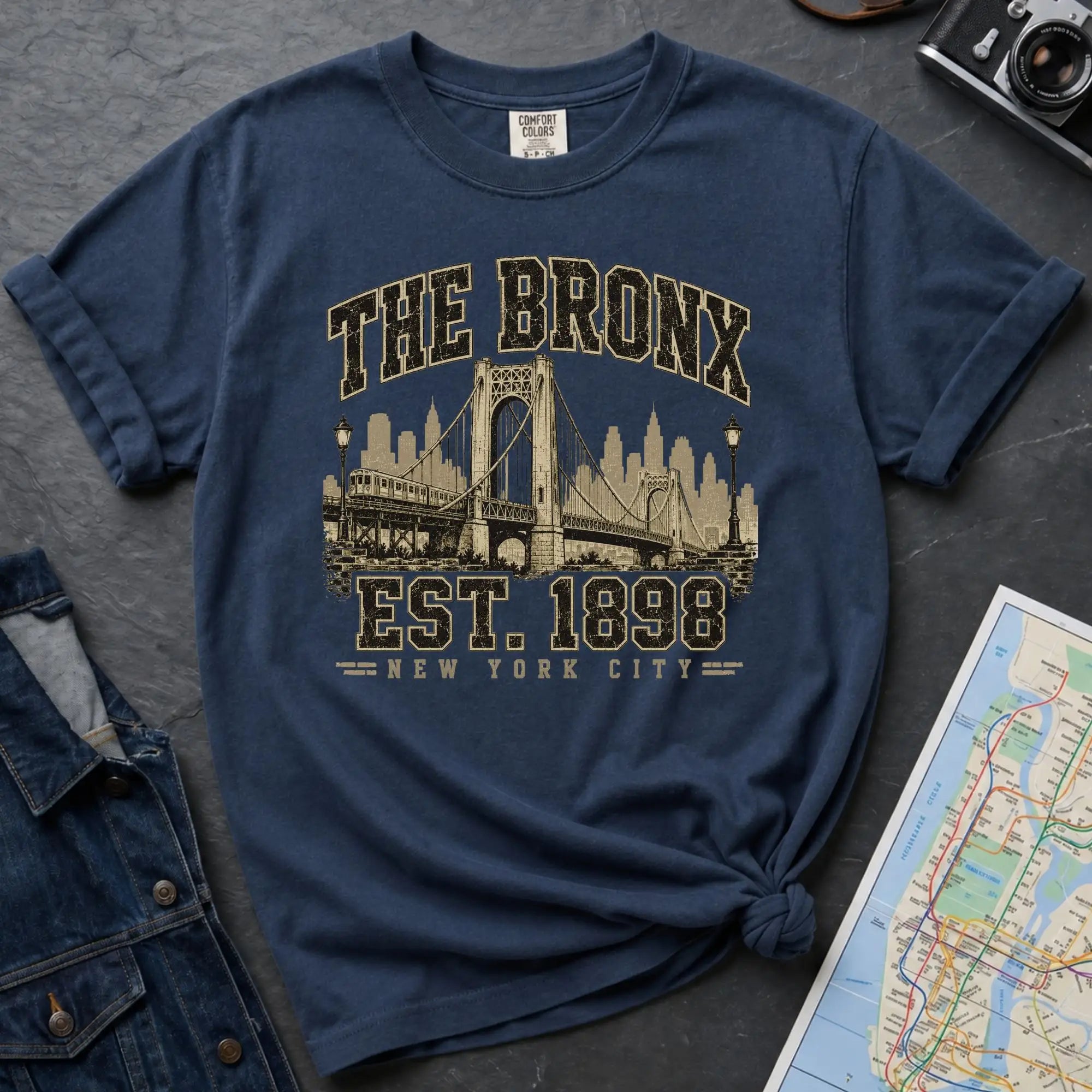 Navy t-shirt with 'The Bronx Est. 1898' graphic on a dark surface with denim jacket and map.