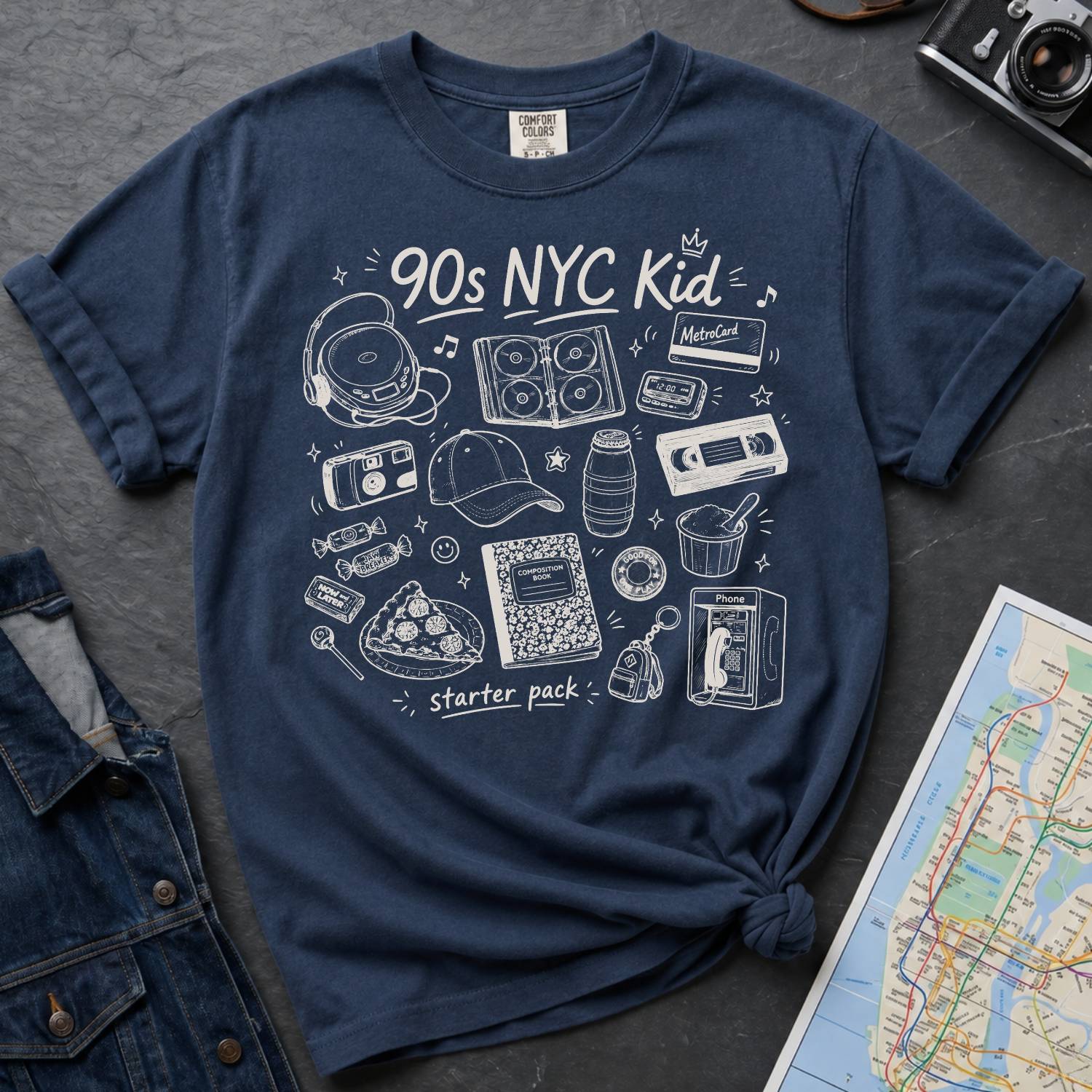 Navy blue Comfort Colors t-shirt with '90s NYC Kid' graphic design on a dark surface with a map and denim jacket.