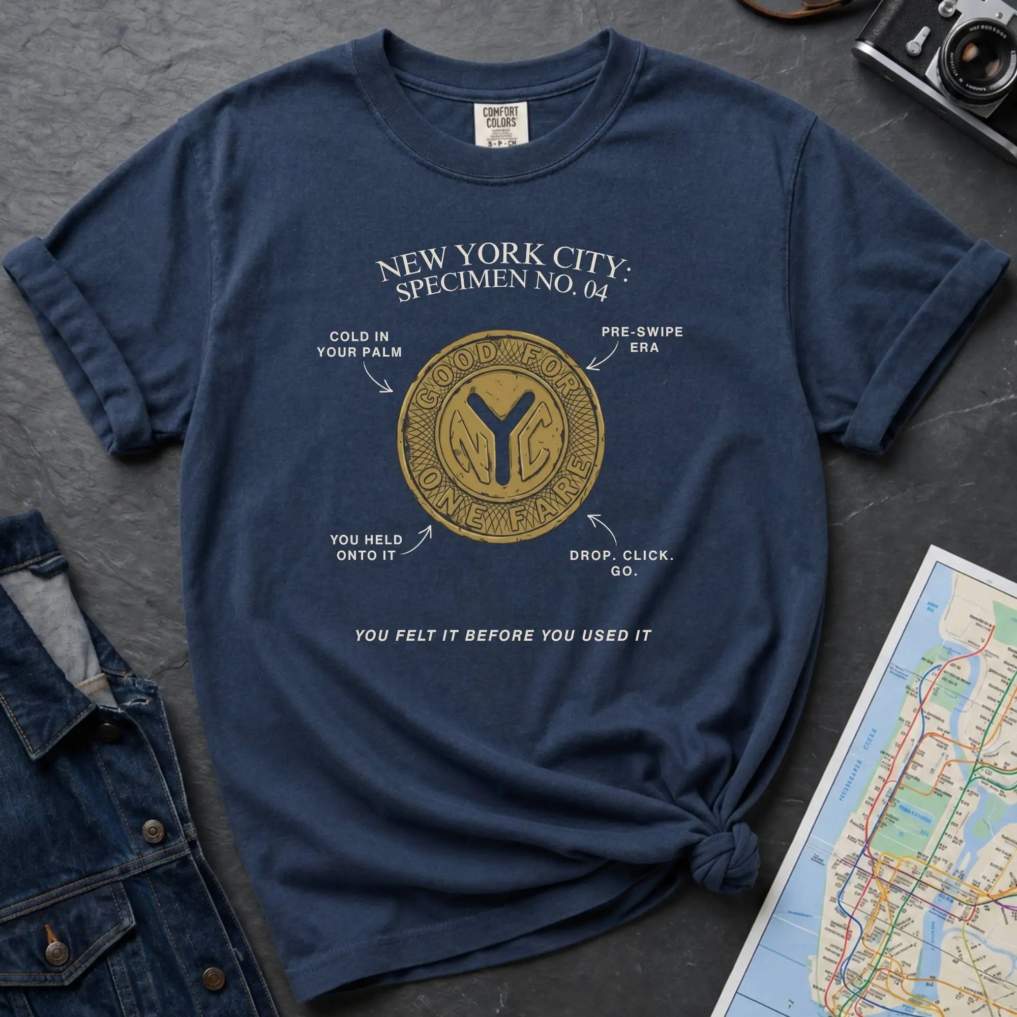 Navy t-shirt with New York City token design on a gray surface with a map and camera.