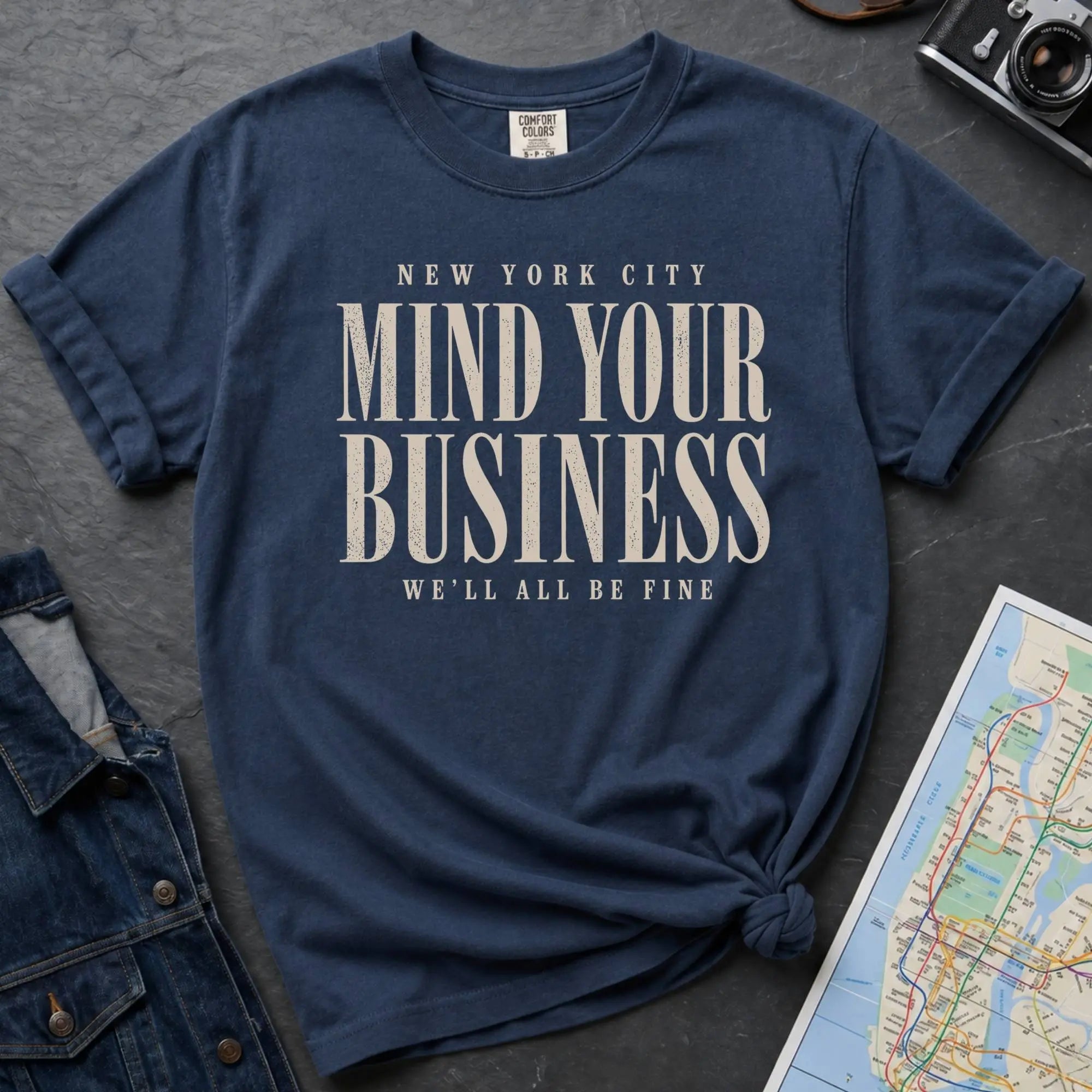 Navy t-shirt with 'New York City Mind Your Business We'll All Be Fine' text on a dark surface with a map and camera.