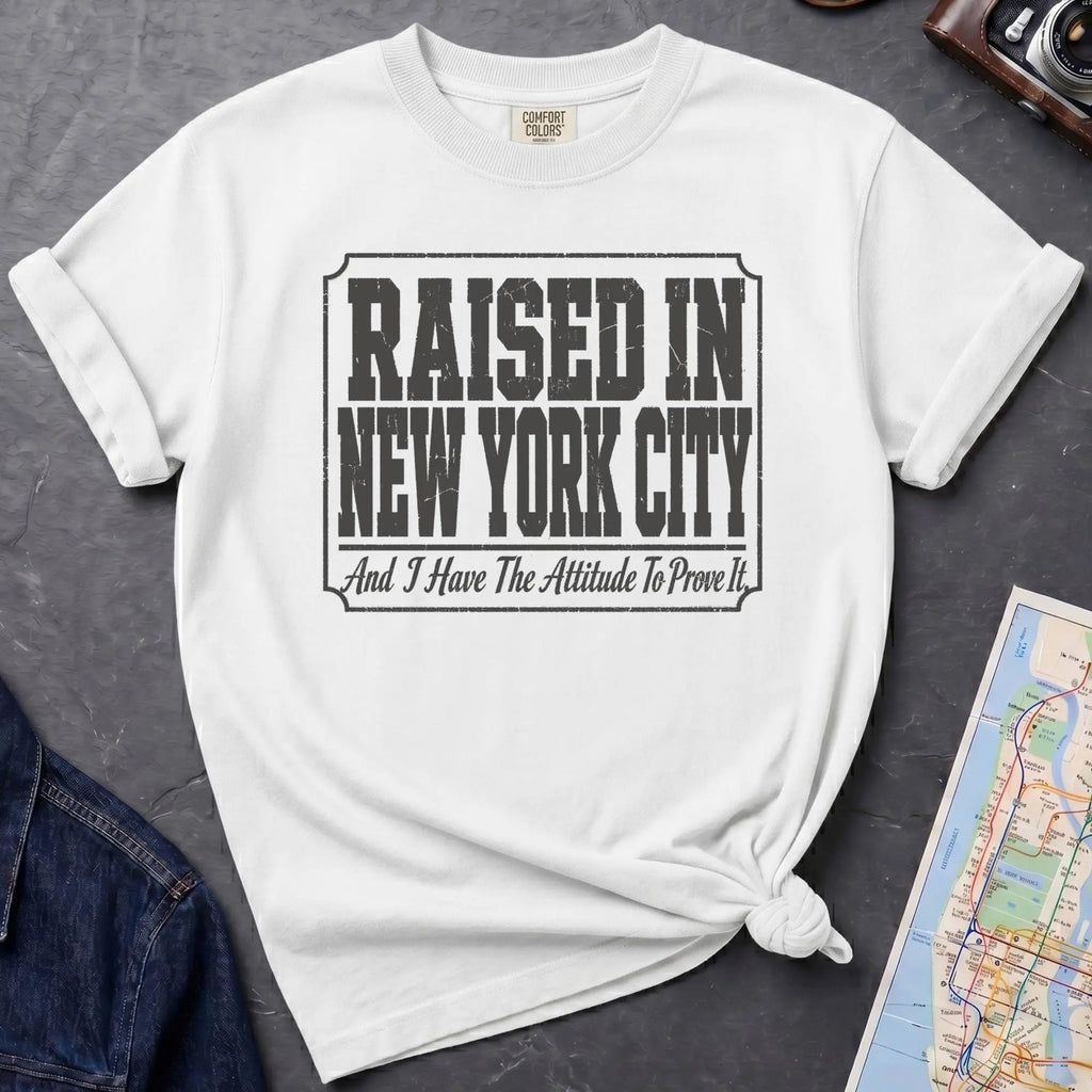 White t-shirt with 'Raised in New York City' text on a dark surface with a jean jacket and a map.