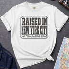 White t-shirt with 'Raised in New York City' text on a dark surface with a jean jacket and a map.