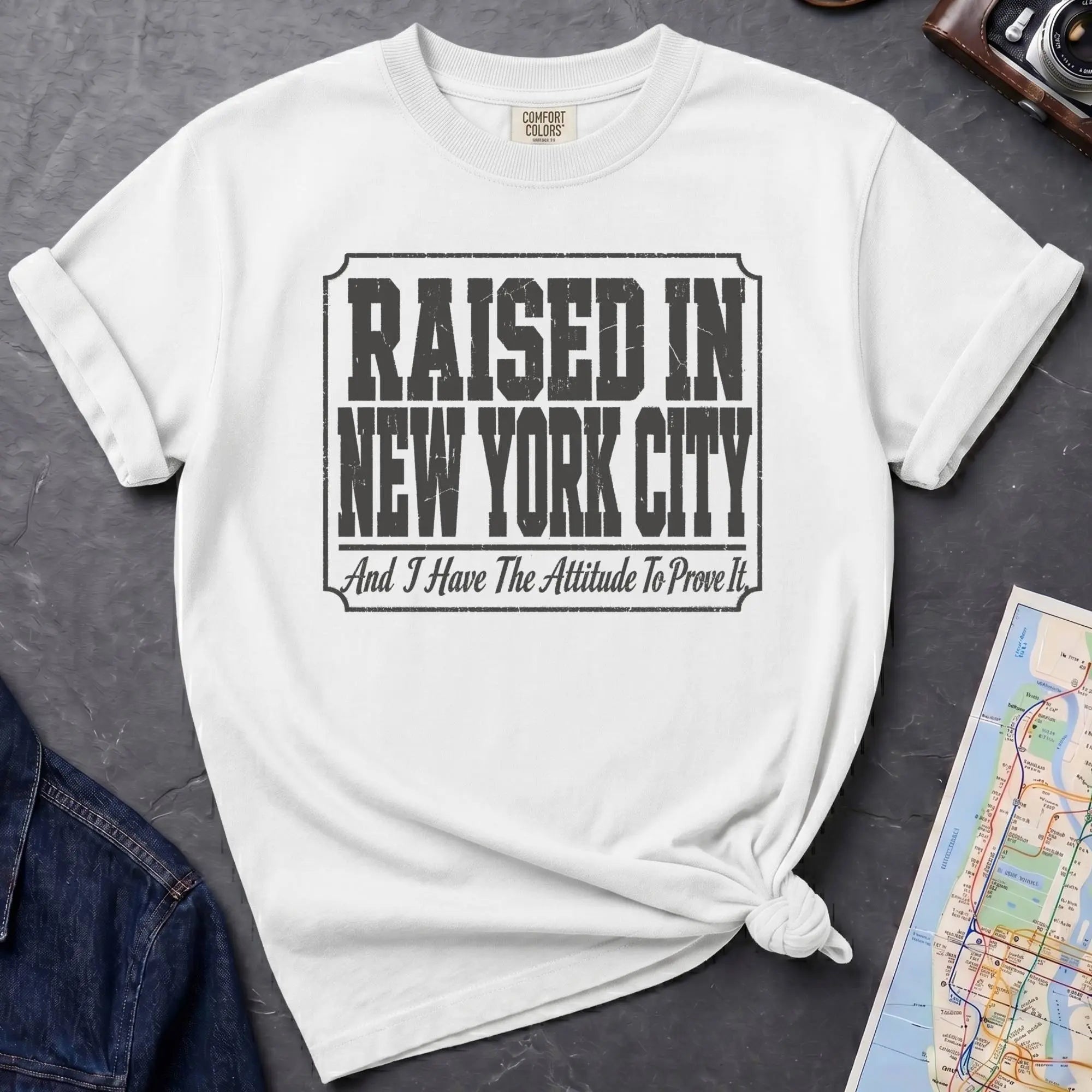 White t-shirt with 'Raised in New York City' text on a dark surface with a jean jacket and a map.