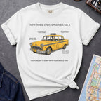 White Comfort Colors t-shirt with yellow taxi graphic and text on a dark surface with denim jacket and map.
