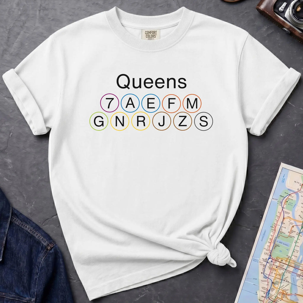 White t-shirt with 'Queens' and subway line numbers on a dark surface with a map and camera.