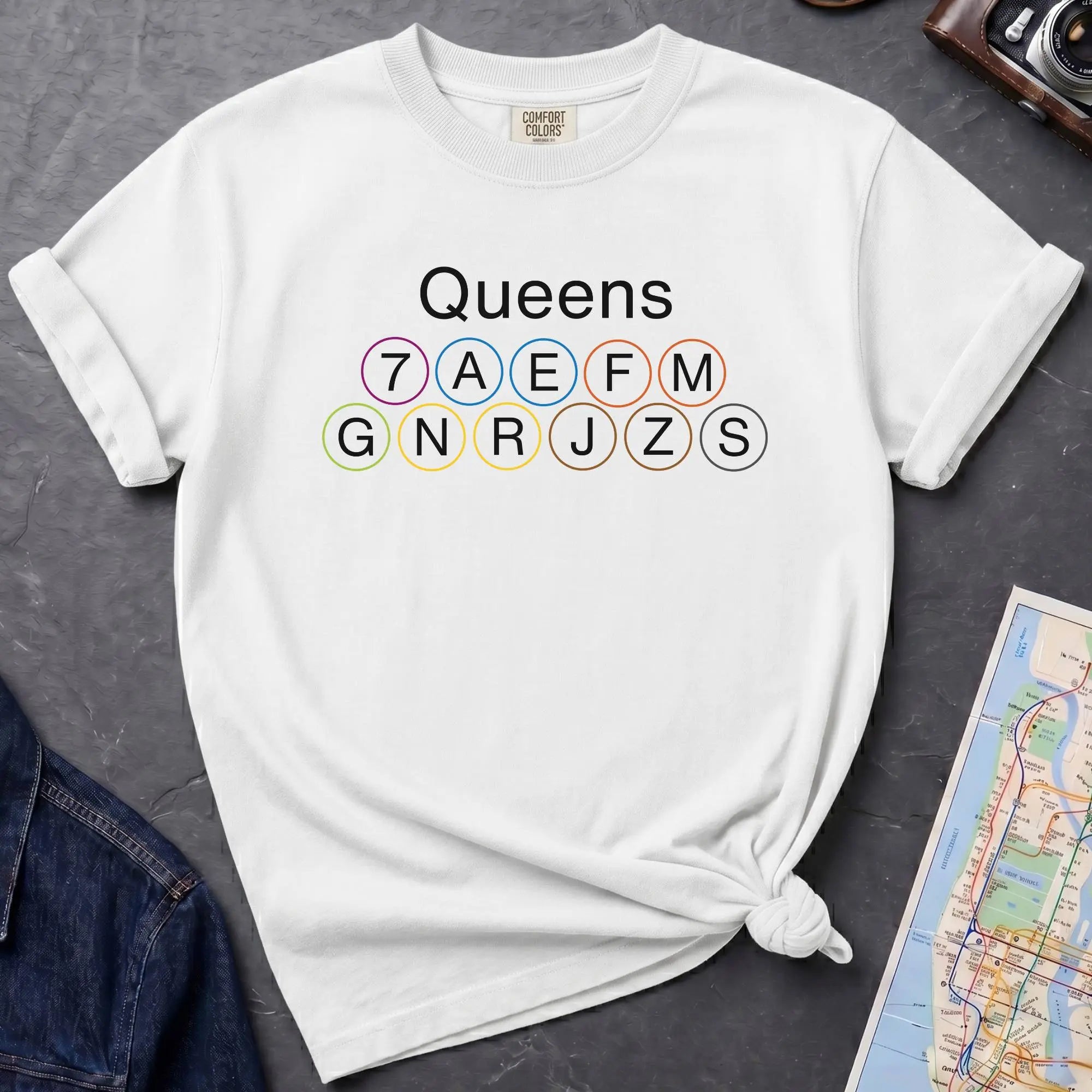 White t-shirt with 'Queens' and subway line numbers on a dark surface with a map and camera.