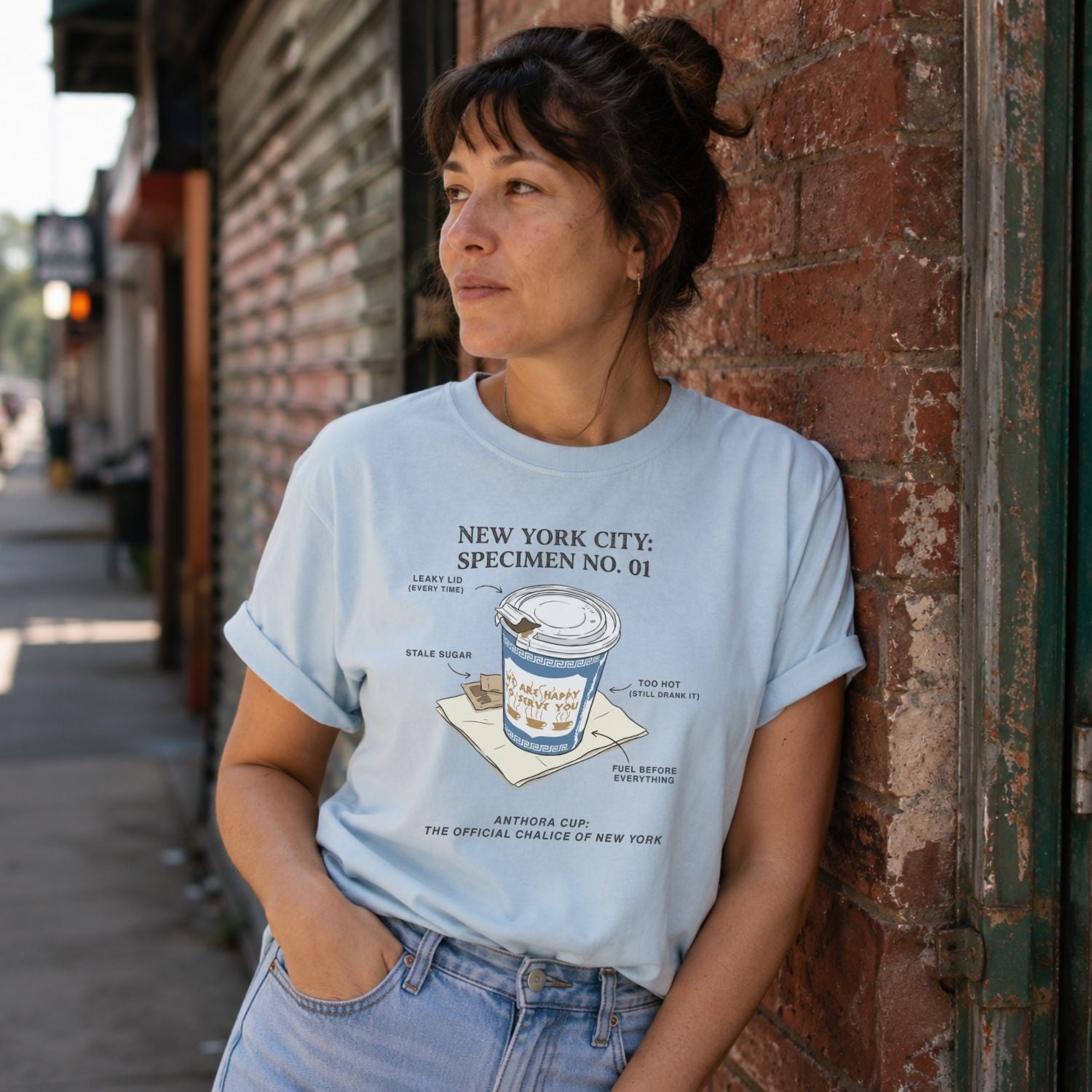 Woman wearing a Light blue Comfort Colors t-shirt featuring a vintage New York City Anthora coffee cup graphic with specimen-style callouts and “The Official Chalice of New York” text, standing against a brick wall.
