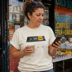 Woman wearing an Ivory Comfort Colors t-shirt with a NYC taxi design, holding a cup and phone, standing in front of a store.