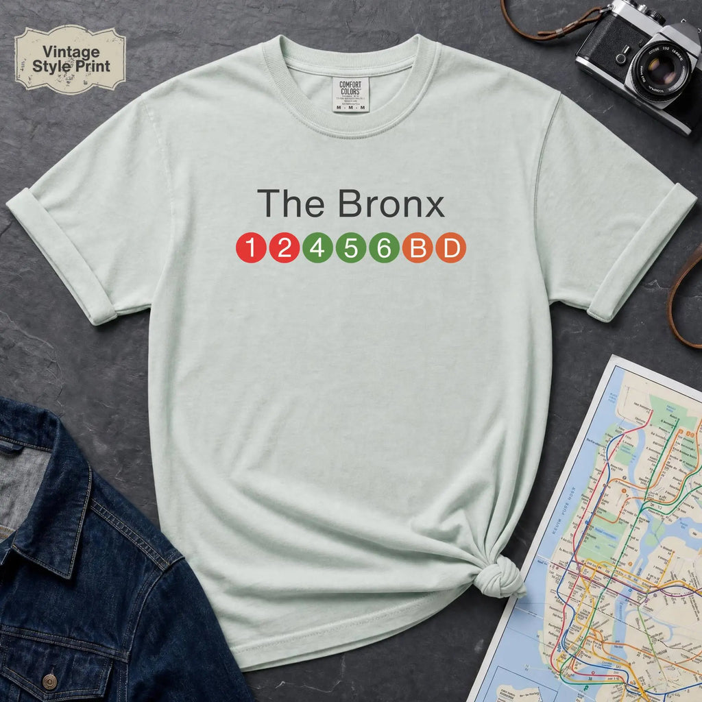 Light blue t-shirt with 'The Bronx' and subway line numbers printed on it, placed on a dark surface with a map and denim jacket.