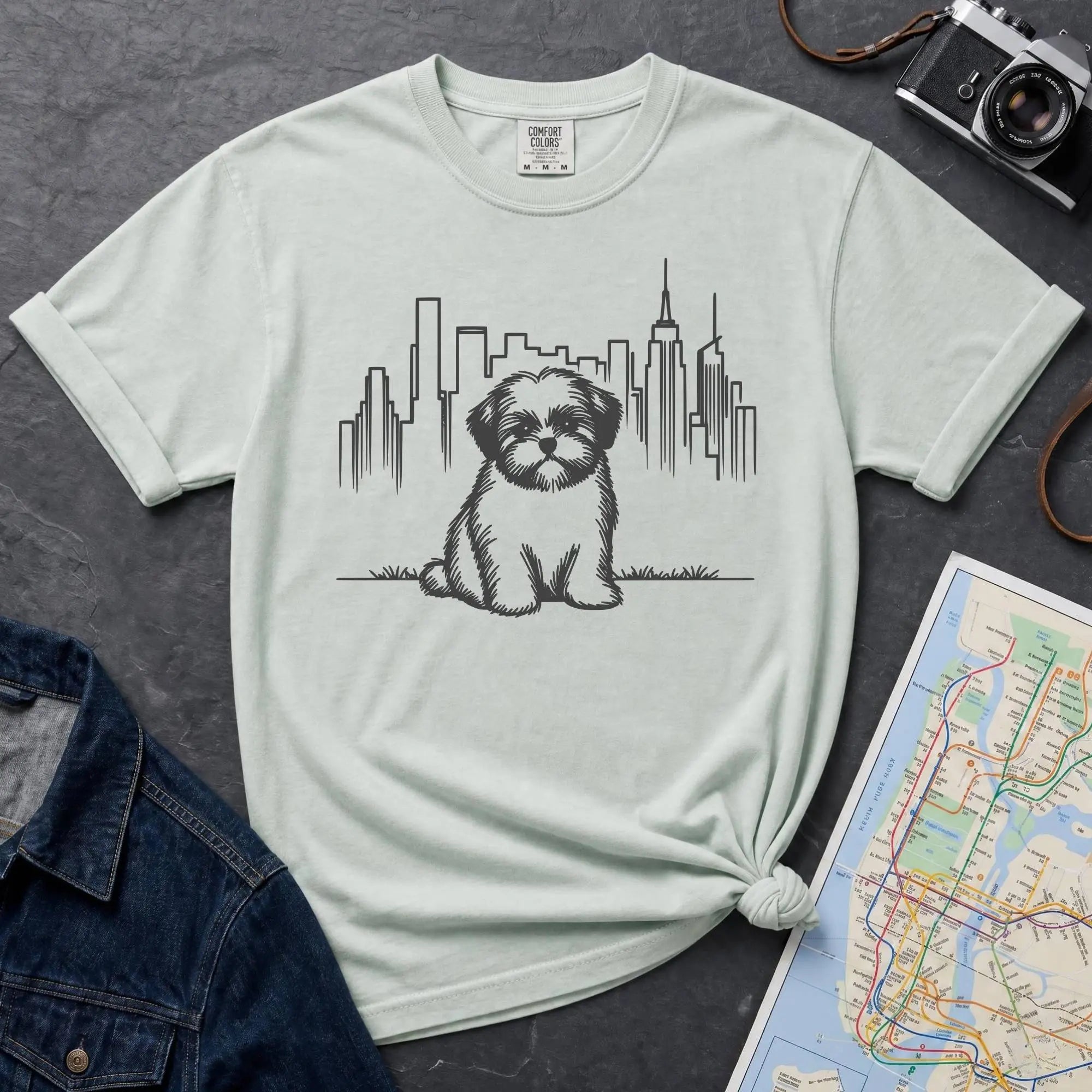 Bay colored t-shirt with a Shih Tzu dog and cityscape design on a dark surface with a map and camera.