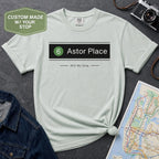 Bay colored t-shirt with '6 Astor Place Still My Stop' text on a textured surface with a map and camera.