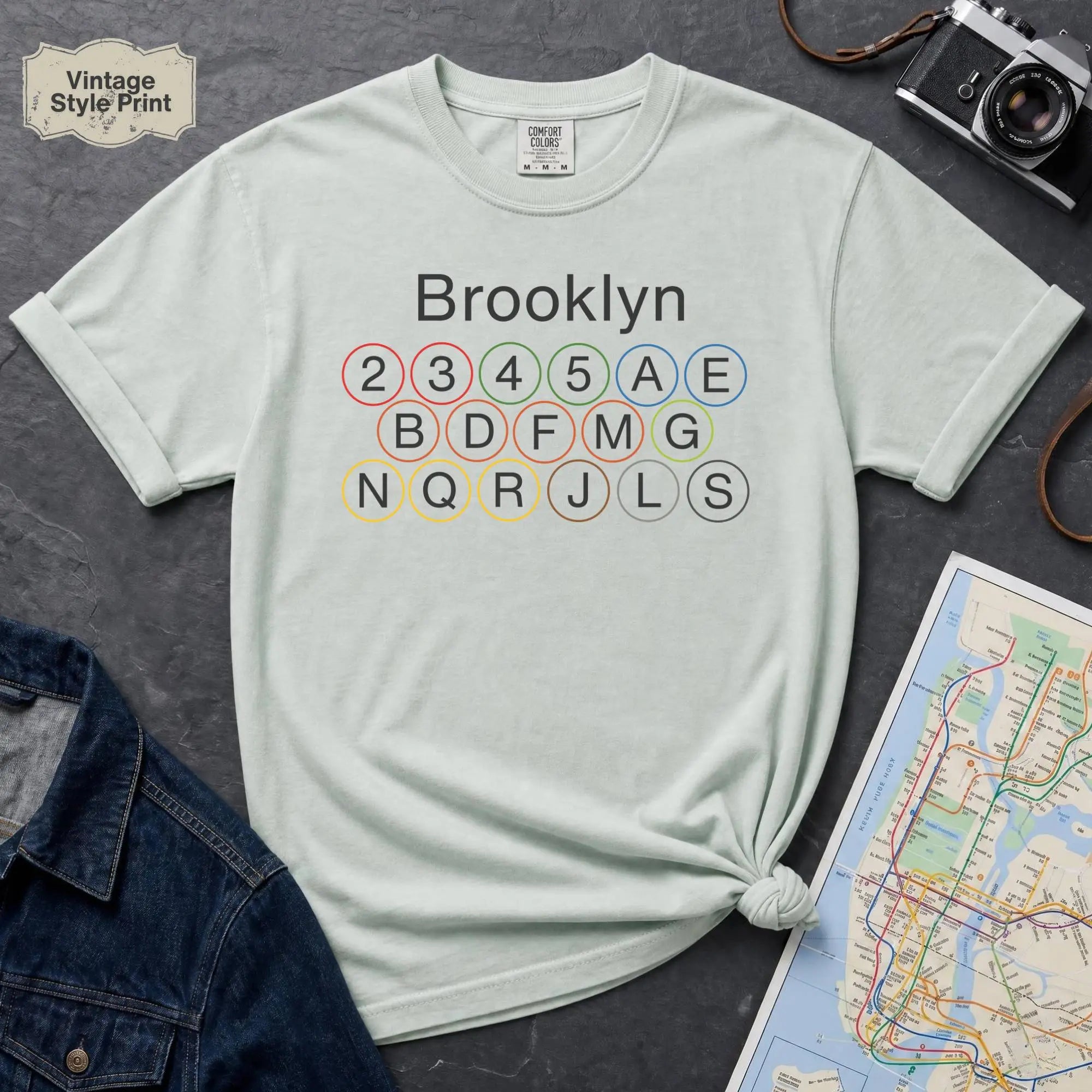 Bay colored t-shirt with 'Brooklyn' and subway line numbers printed on it, placed on a dark surface with a map and denim jacket.