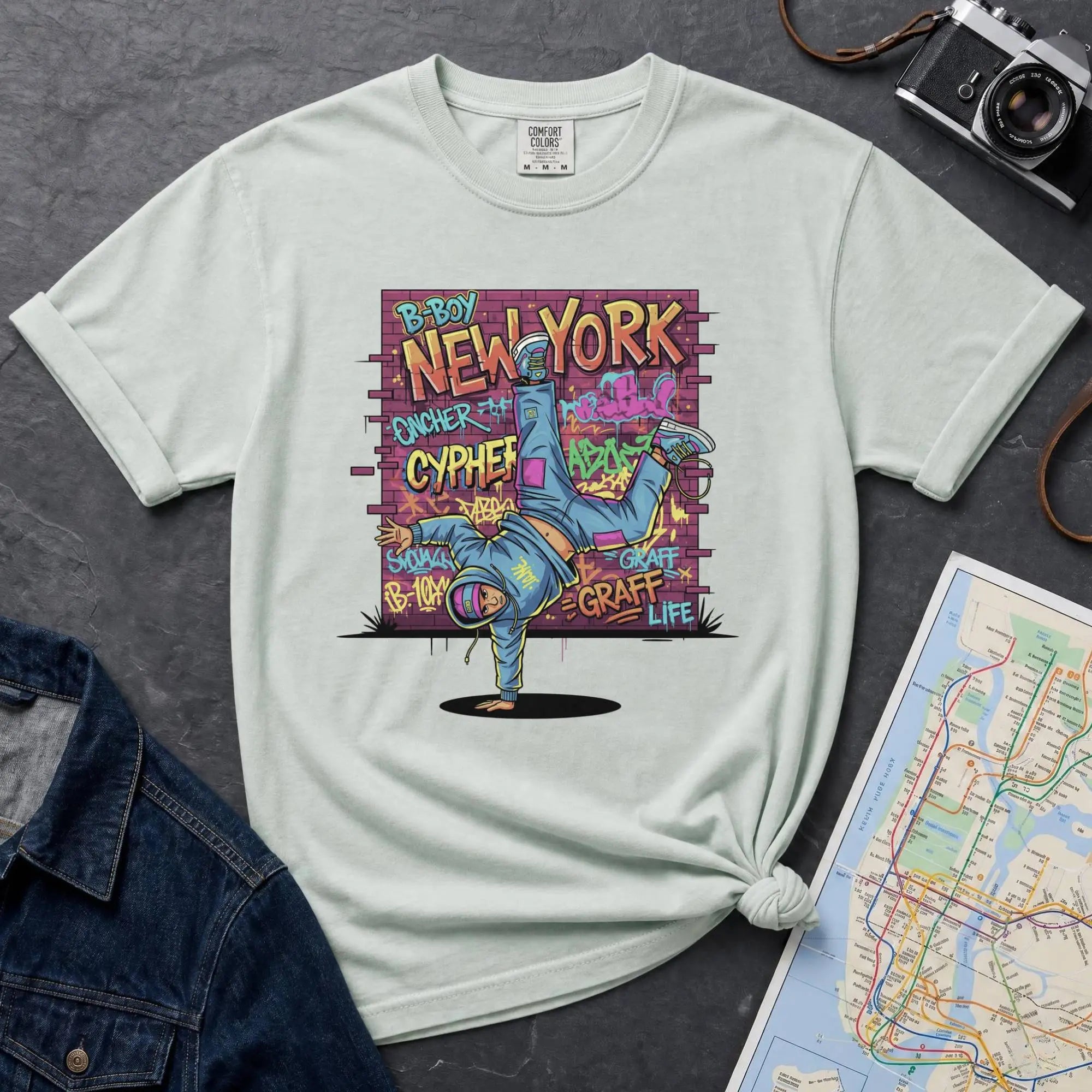 Bay colored t-shirt with a colorful graphic design featuring a breakdancer and text on a dark surface with a denim jacket and a map.