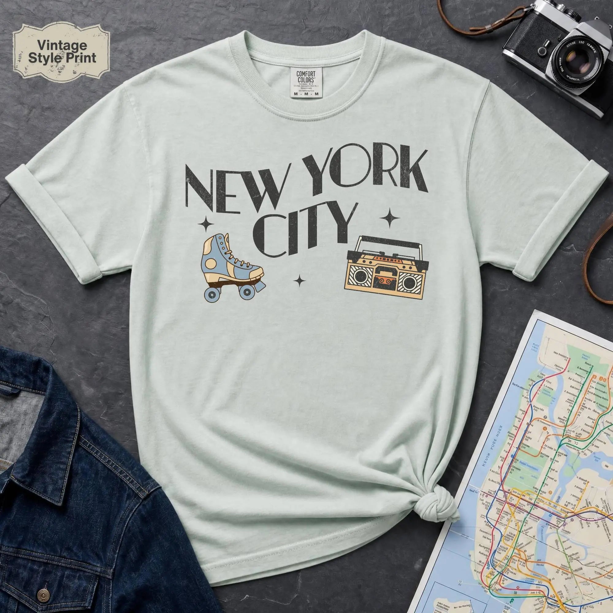 Bay colored t-shirt with 'New York City' text and a roller skate and boom box on a dark surface with a map and camera.