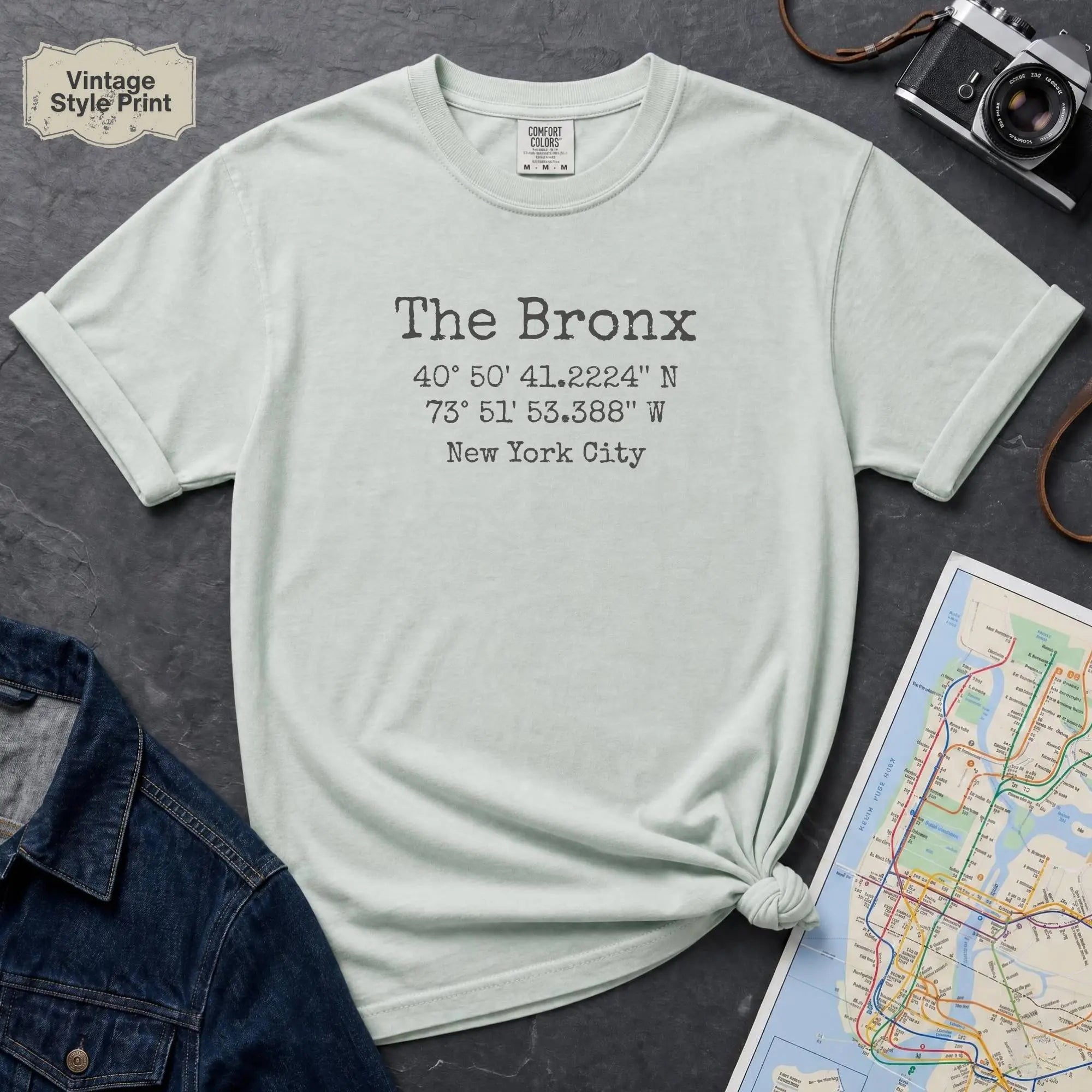 Bay colored t-shirt with 'The Bronx' text and coordinates, placed on a dark surface with a map and denim jacket.