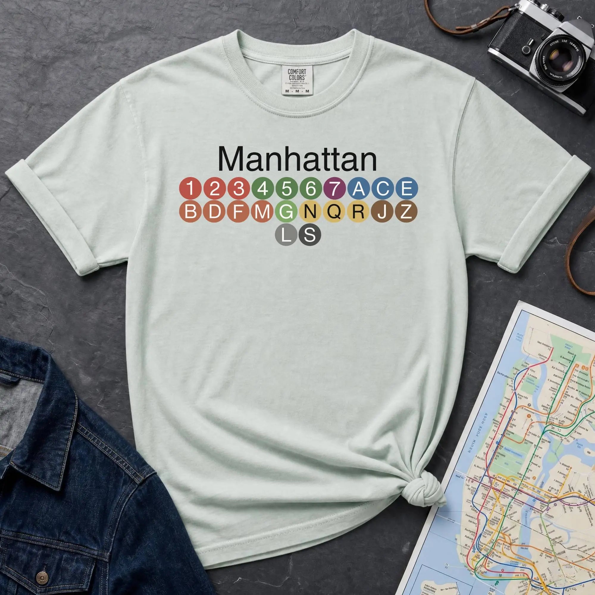 Bay colored Comfort Colors t-shirt featuring a minimalist Manhattan subway line graphic with colorful NYC train icons.