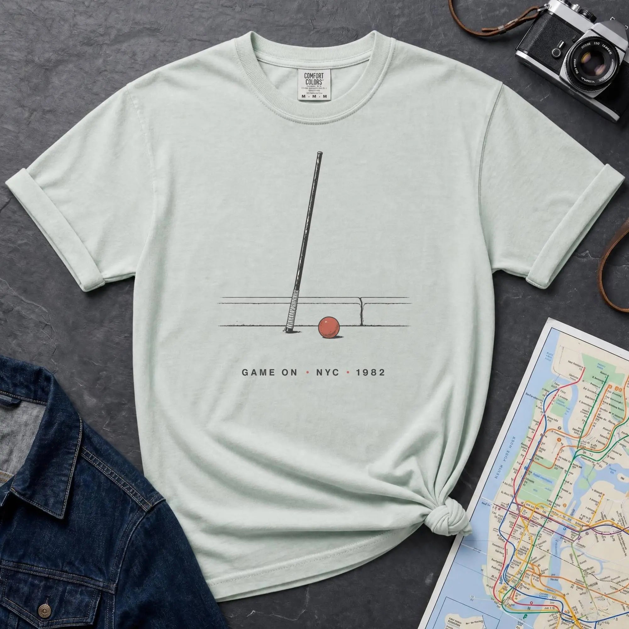 Bay colored Comfort Colors t-shirt featuring a minimalist stickball bat and ball graphic with “Game On • NYC • 1982” text.