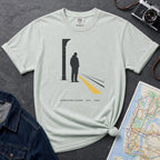 Bay colored t-shirt with a graphic design of a person on a train platform, placed on a dark surface with a map and denim jacket.