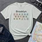 Bay colored t-shirt with 'Brooklyn' and subway line numbers printed on it, placed on a dark surface with a map and denim jacket.
