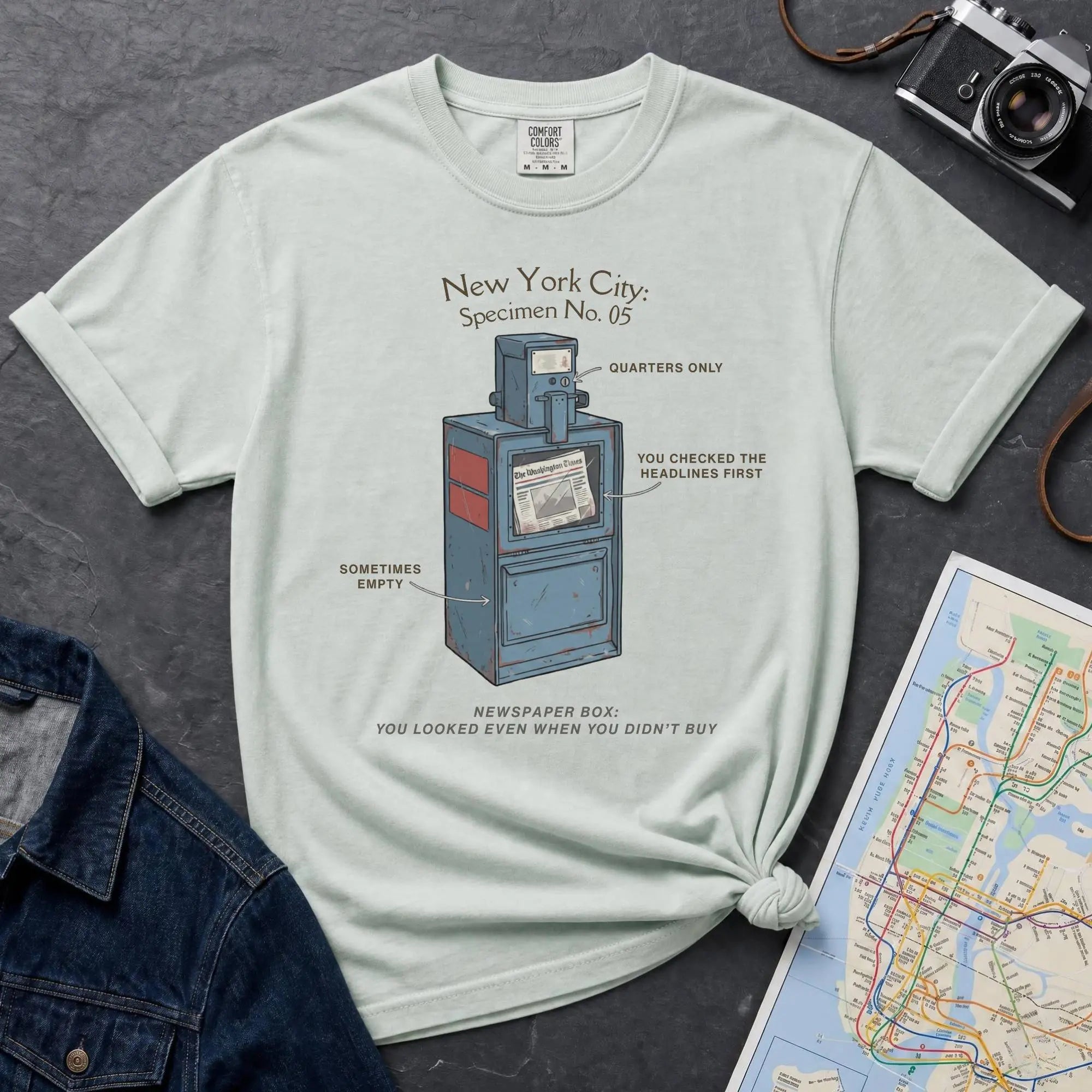 Bay colored t-shirt with a newspaper box graphic and text on a textured surface with a camera and map.