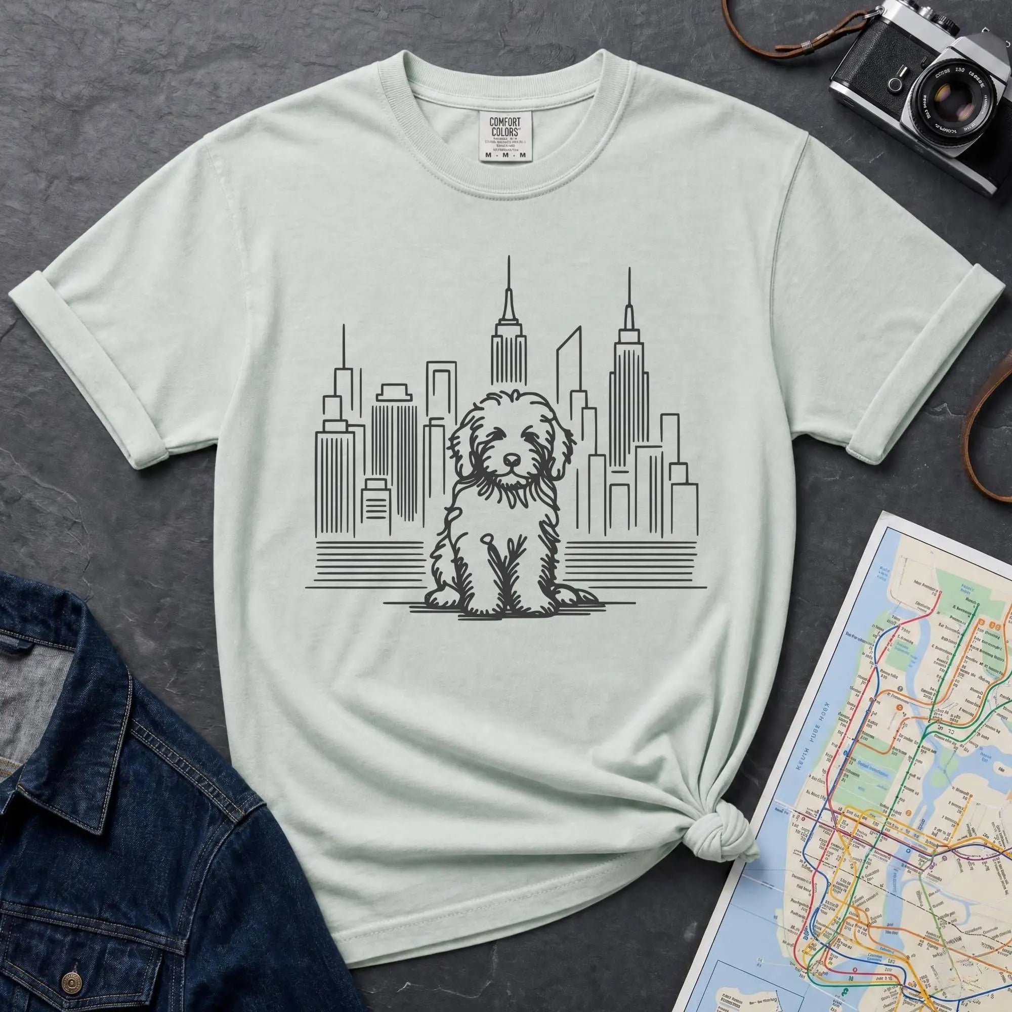 Bay colored t-shirt with a Golden Doodle dog and cityscape design on a gray surface with a map and camera.