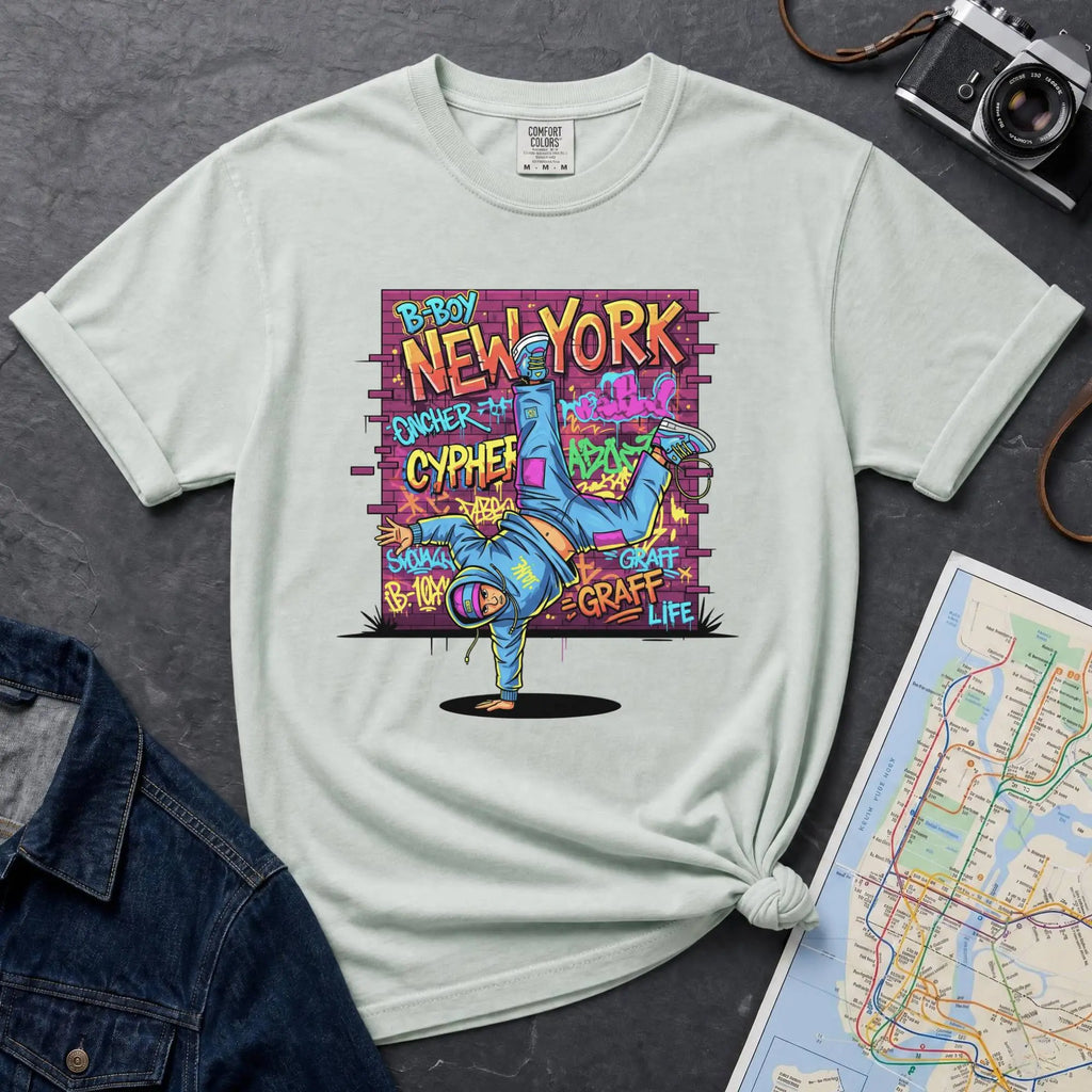 Bay colored t-shirt with a colorful graphic design featuring a breakdancer and text on a dark surface with a denim jacket and a map.