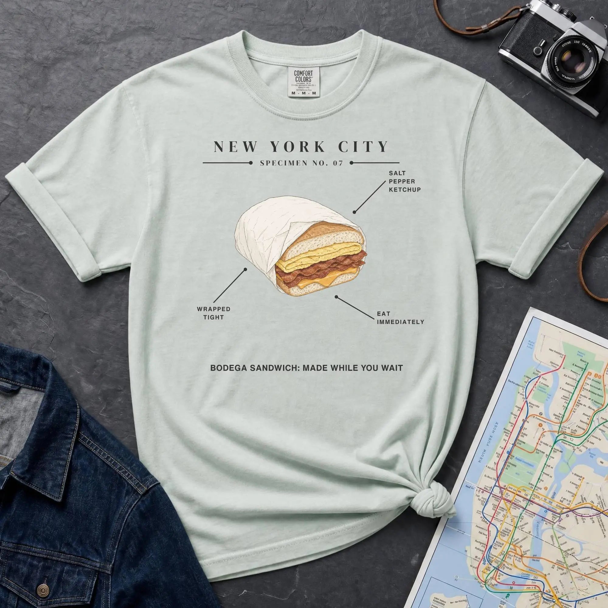 Bay colored Comfort Colors t-shirt featuring a New York City bodega sandwich graphic with specimen-style callouts and “Bodega Sandwich: Made While You Wait” text.