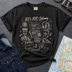 Black t-shirt with '80s NYC Subway Survival Kit' graphic design on a dark surface with a map and denim jacket.