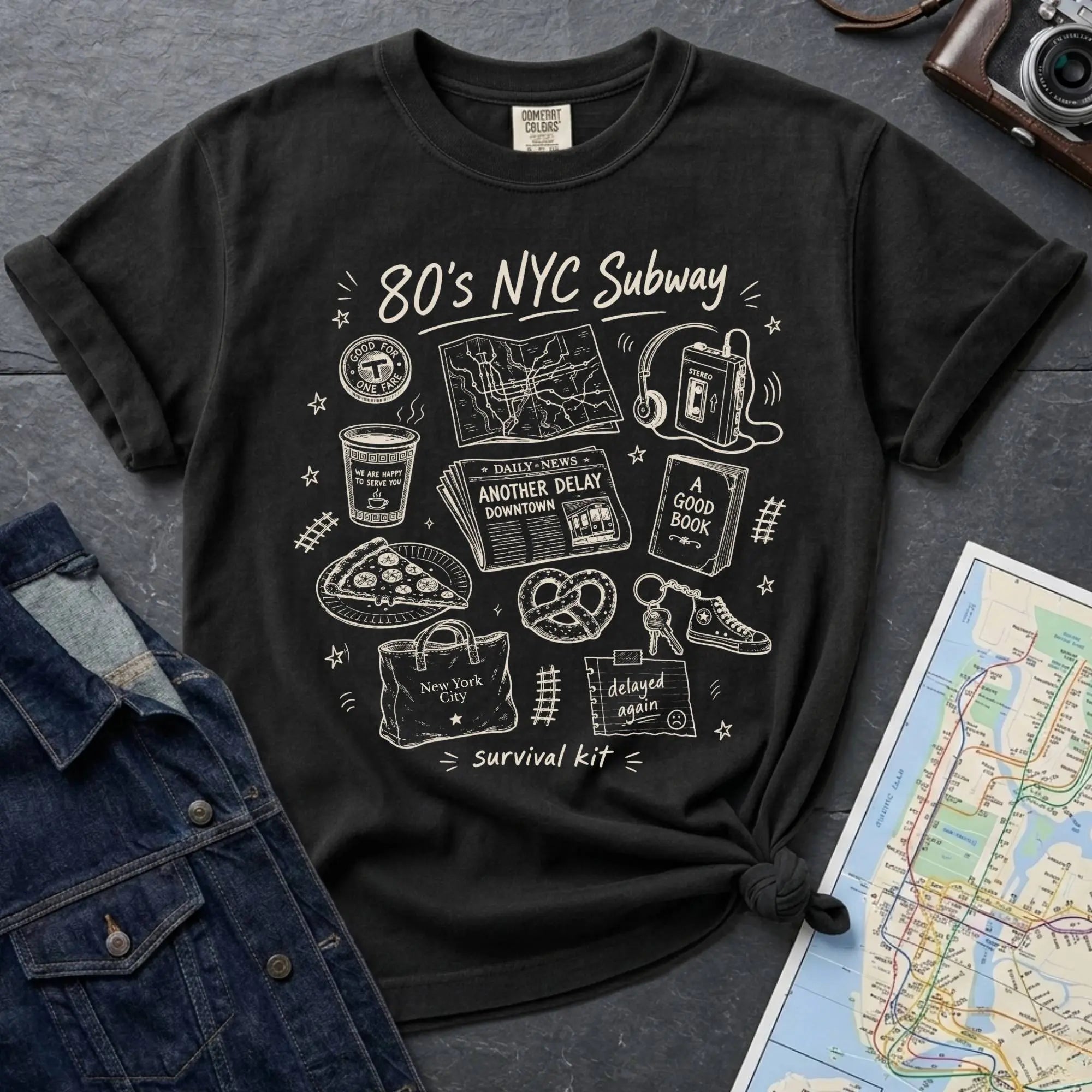 Black t-shirt with '80s NYC Subway Survival Kit' graphic design on a dark surface with a map and denim jacket.