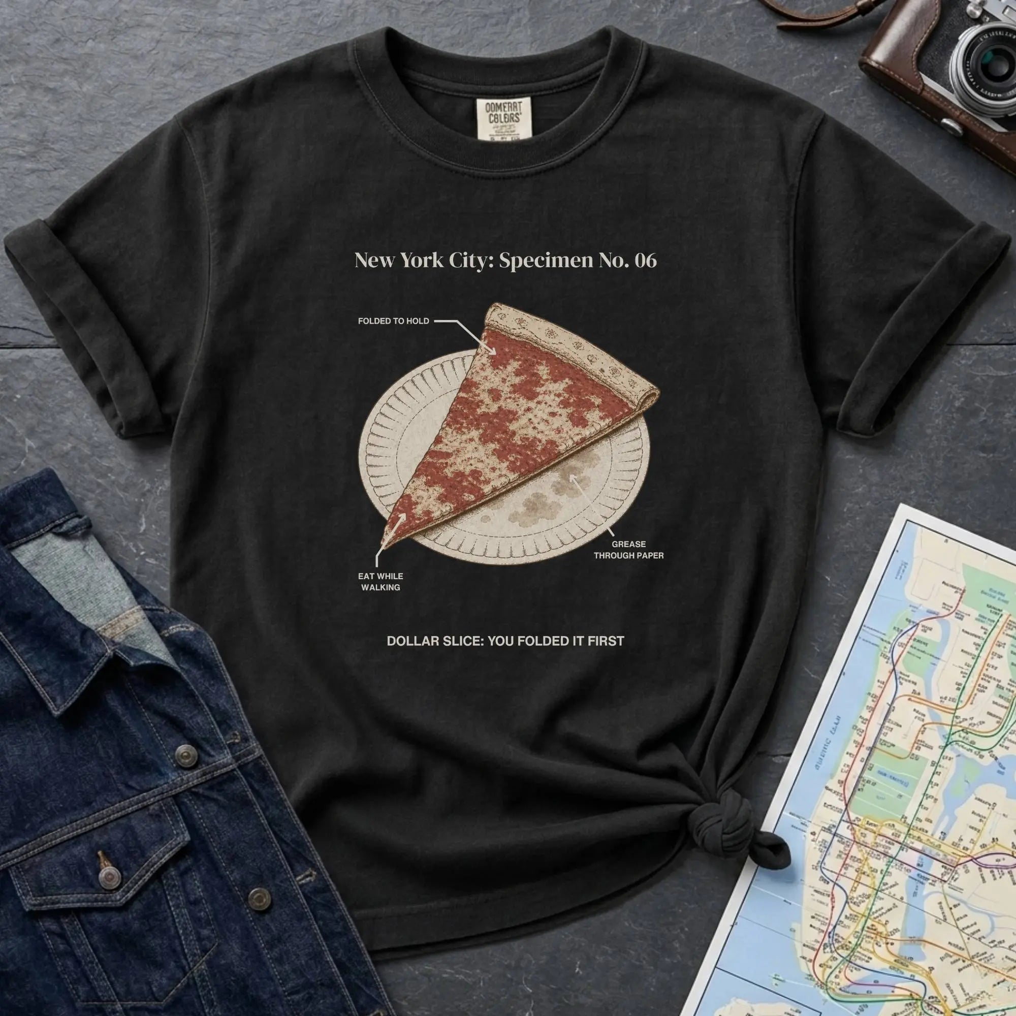 Black Comfort Colors t-shirt featuring a vintage New York City dollar slice pizza graphic with specimen-style callouts and a folded slice on a paper plate.