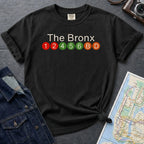 Black Comfort Colors t-shirt featuring a minimalist Bronx subway line graphic with The Bronx text and colorful NYC train icons.