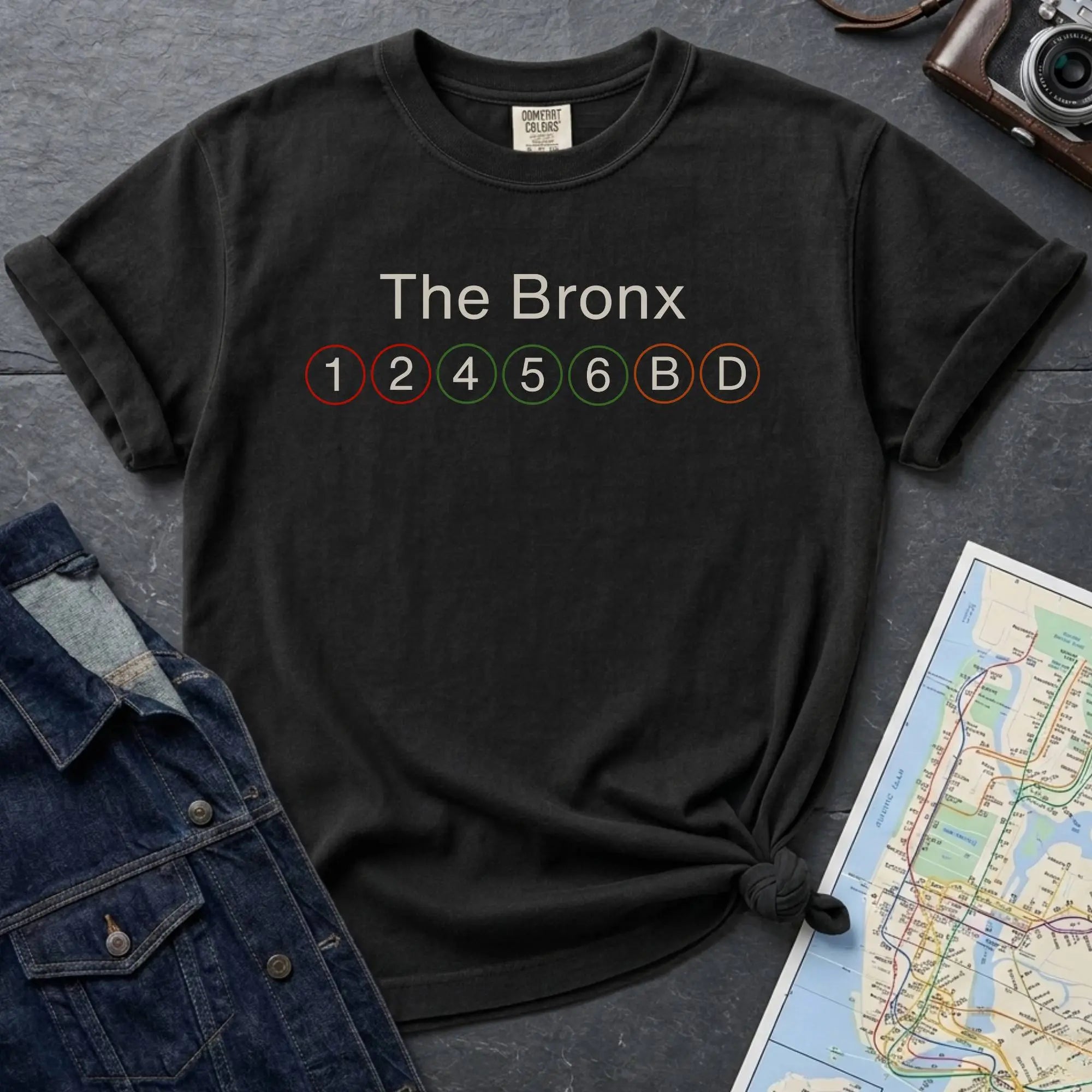 Black t-shirt with 'The Bronx' and subway line numbers on a dark surface with a map and camera.
