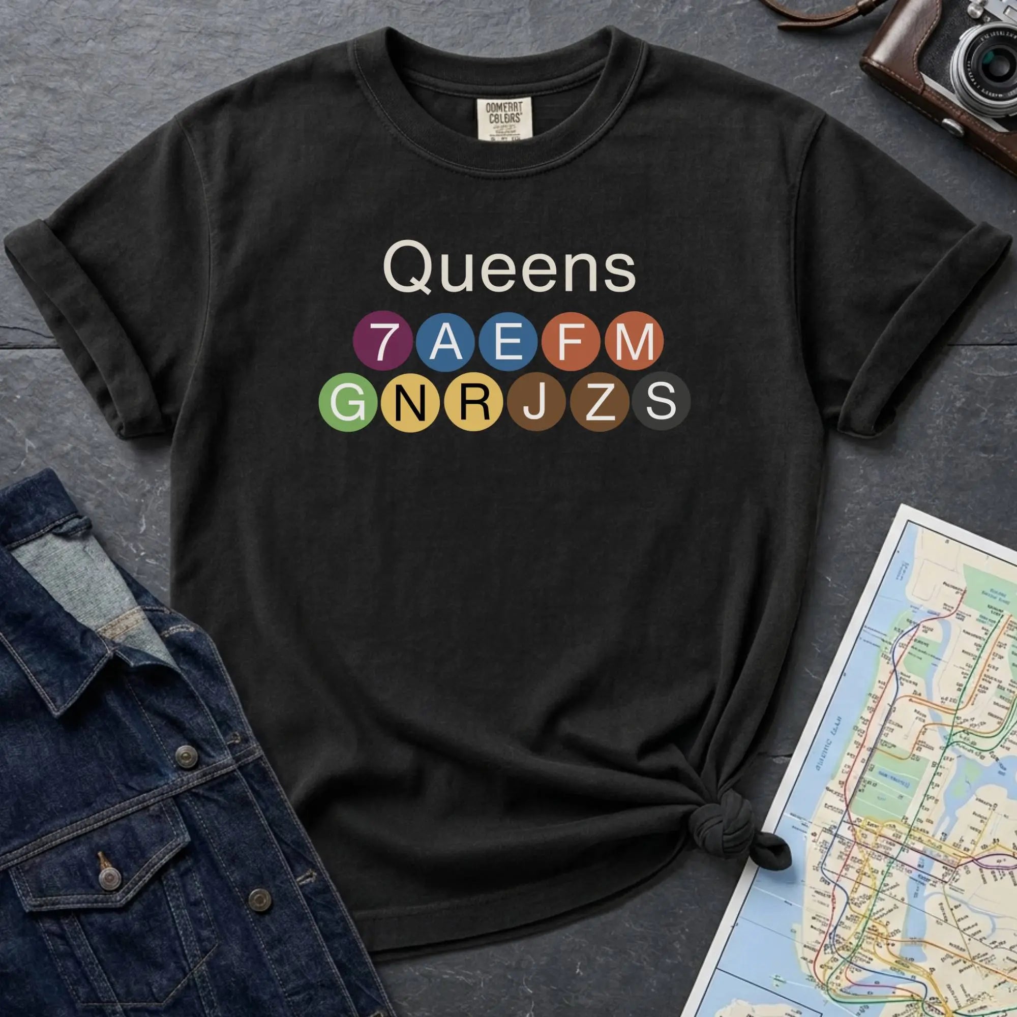 Black Black Comfort Colors t-shirt featuring a minimalist Queens subway line graphic with colorful NYC train icons.