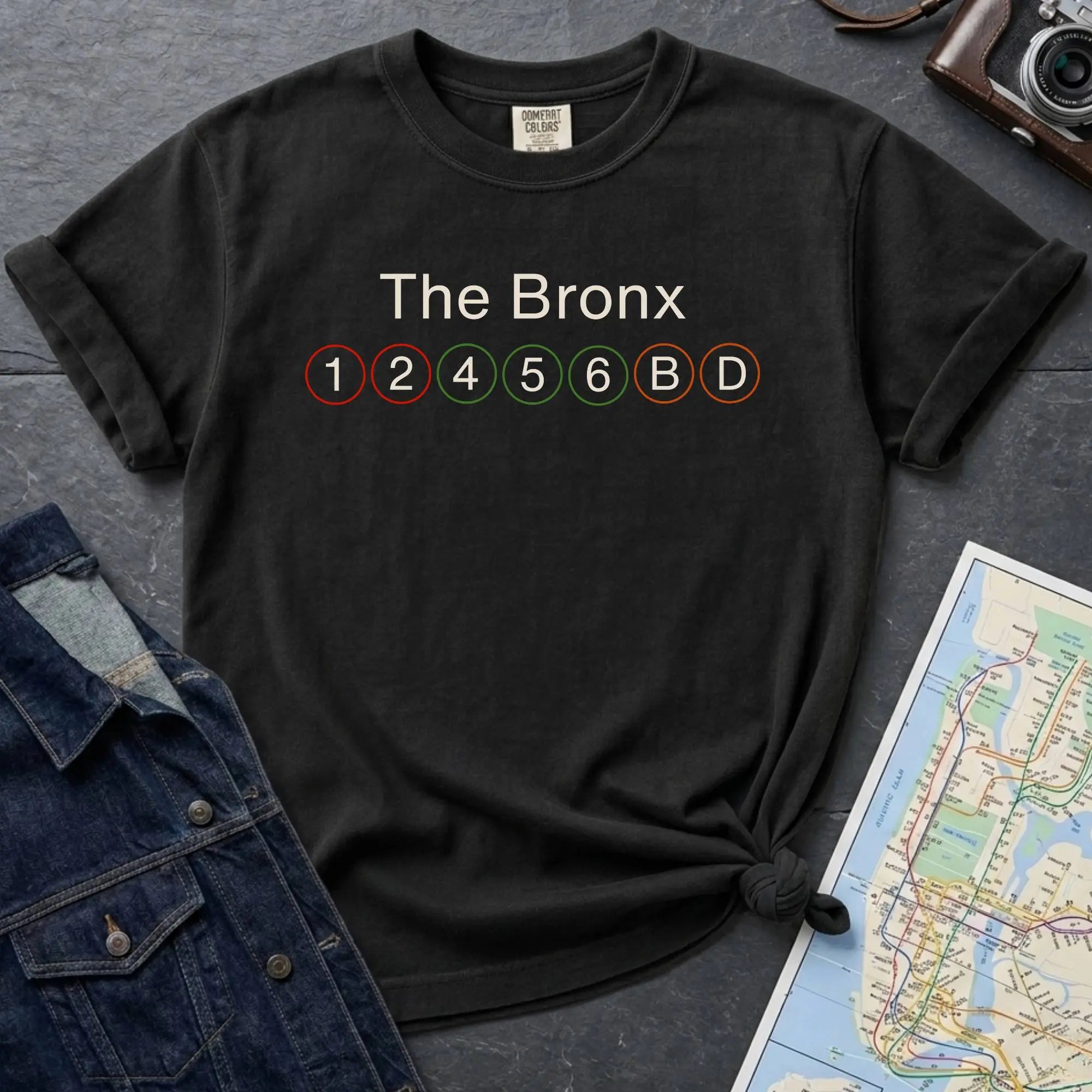 Black t-shirt with 'The Bronx' and subway line numbers on a dark surface with a map and camera.