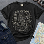 Black t-shirt with '80s NYC Subway Survival Kit' graphic design on a dark surface with a map and denim jacket.