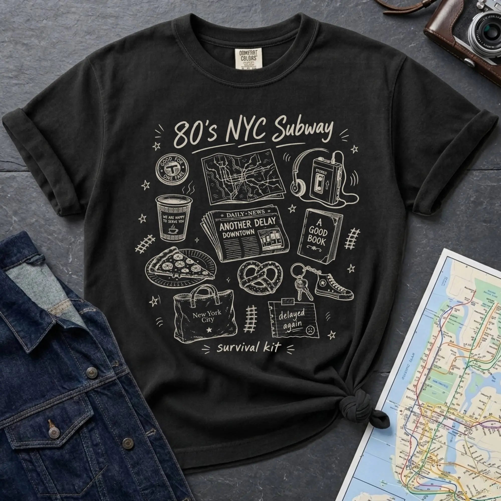 Black t-shirt with '80s NYC Subway Survival Kit' graphic design on a dark surface with a map and denim jacket.