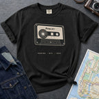 Black t-shirt with cassette tape graphic and text on a gray surface with denim jacket and map.