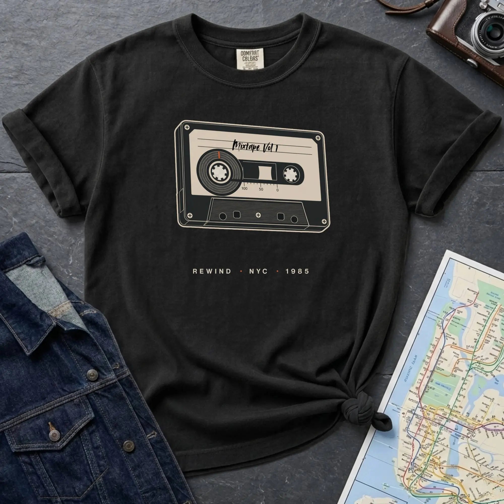 Black t-shirt with cassette tape graphic and text on a gray surface with denim jacket and map.