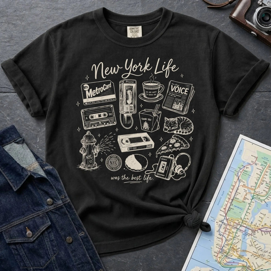 Black Comfort Colors t-shirt featuring a New York Life Was the Best Life graphic with hand-drawn NYC nostalgia icons including a MetroCard, cassette tape, pay phone, coffee cup, newspaper, pizza slice, fire hydrant, Walkman, and bodega cat.