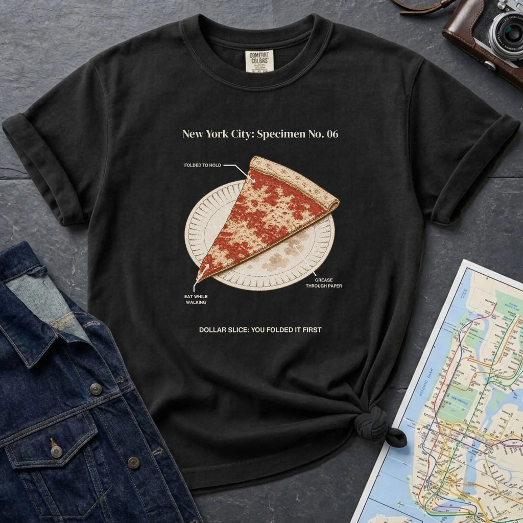 Black Comfort Colors t-shirt featuring a vintage New York City dollar slice pizza graphic with specimen-style callouts and a folded slice on a paper plate.