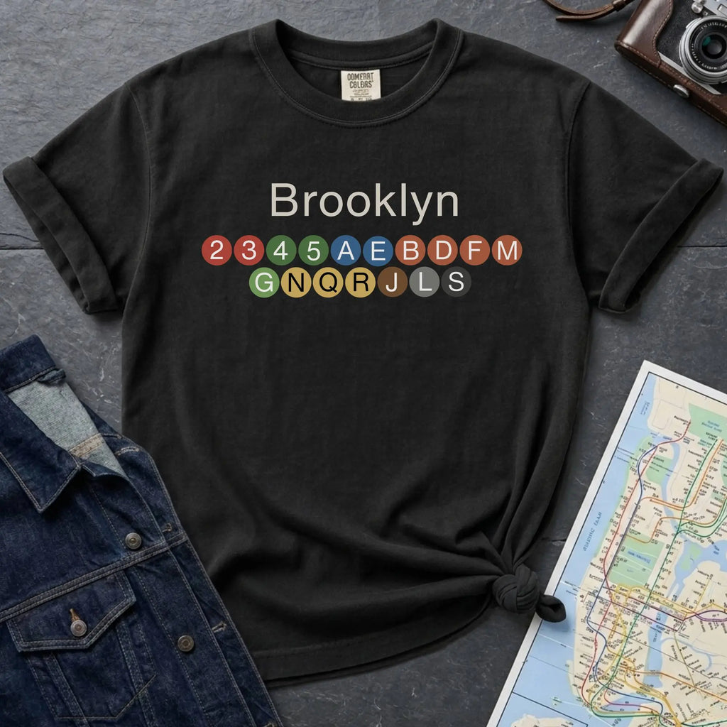 Black t-shirt with 'Brooklyn' and subway line numbers on a dark surface with a map and camera.
