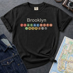 Black t-shirt with 'Brooklyn' and subway line numbers on a dark surface with a map and camera.