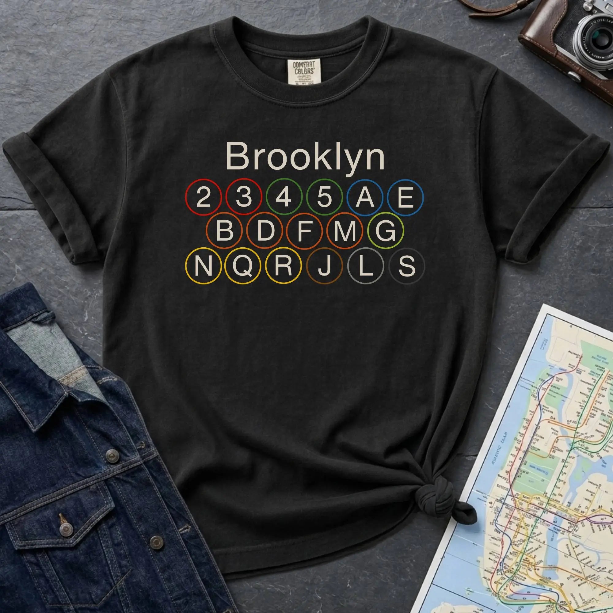 Black Comfort Colors t-shirt featuring the word “Brooklyn” above colorful NYC subway line circles representing the subway routes that run through Brooklyn.