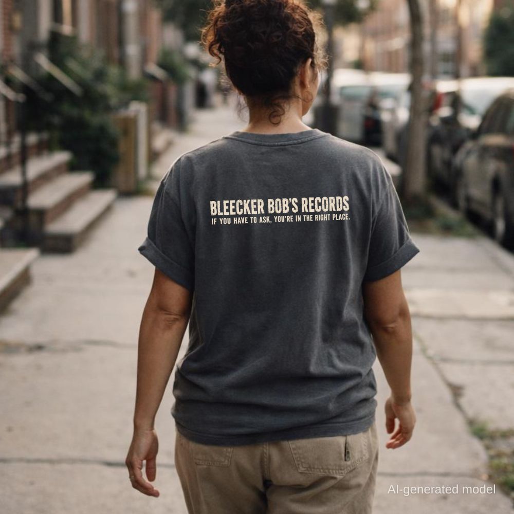 A woman wearing a pepper-colored Comfort Colors T-shirt with ivory Bleecker Bob’s Records NYC back print, while walking down a city street.
