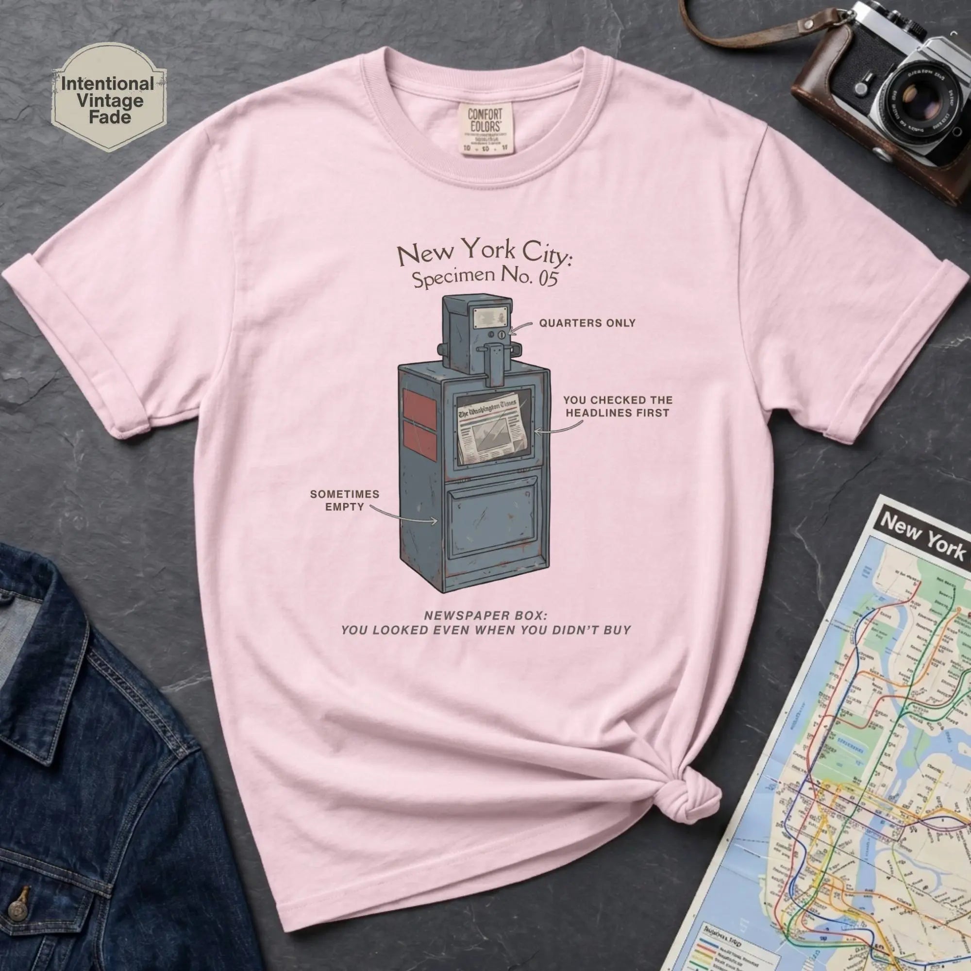 Pink t-shirt with a graphic design of a vintage newspaper box and text on a gray surface with a map and camera.
