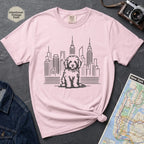 Pink t-shirt with a Golden Doodle dog and cityscape design on a gray surface with a map and camera.