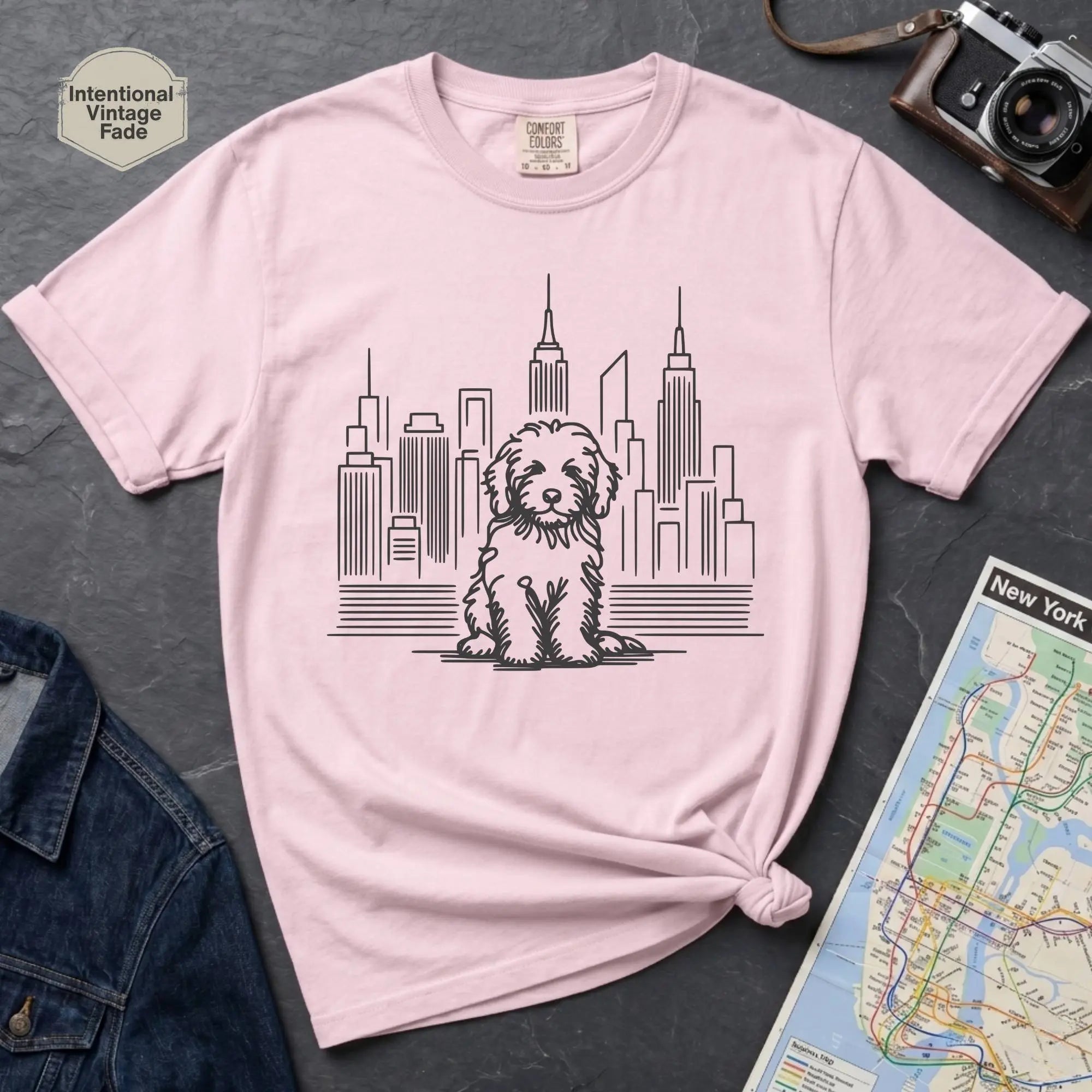Pink t-shirt with a Golden Doodle dog and cityscape design on a gray surface with a map and camera.