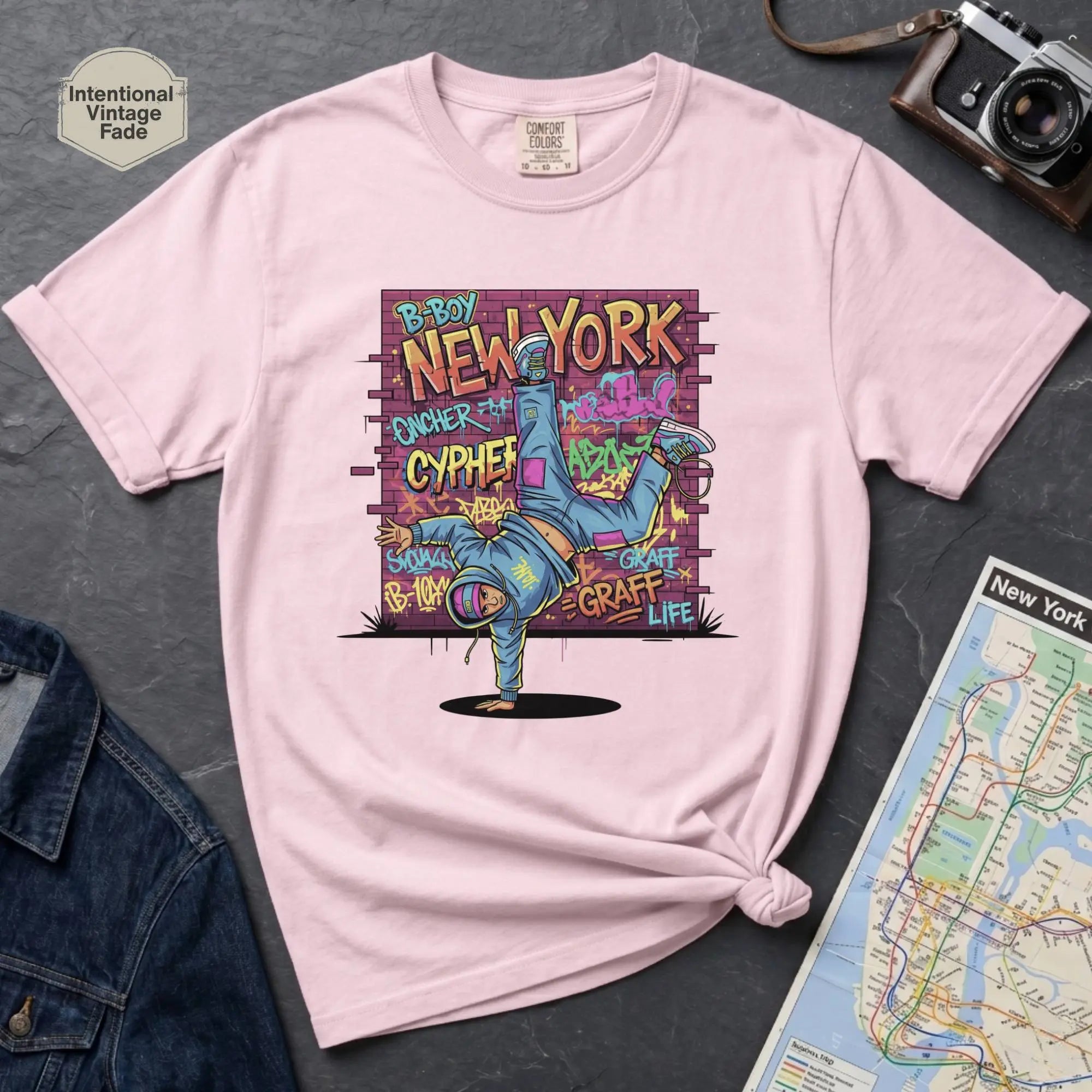 Pink t-shirt with a colorful graphic design featuring a breakdancer and text on a dark surface with a denim jacket and a map.