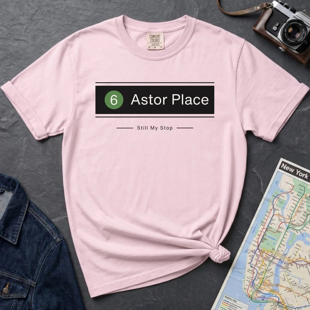 Pink t-shirt with 'Astor Place' design on a dark surface with a map and camera.