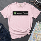 Pink t-shirt with 'Astor Place' design on a dark surface with a map and camera.
